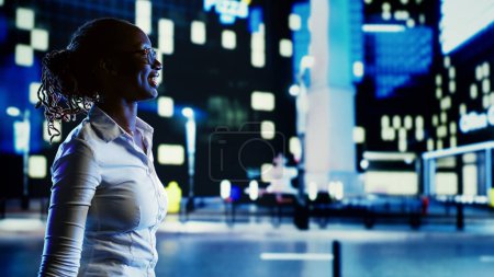 Téléchargez les photos : Femme afro-américaine souriante errant autour des boulevards de la ville pendant la nuit, pointant vers des publicités intéressantes OOH sur les bâtiments. Femme d'affaires flânant dans les rues vides la nuit - en image libre de droit