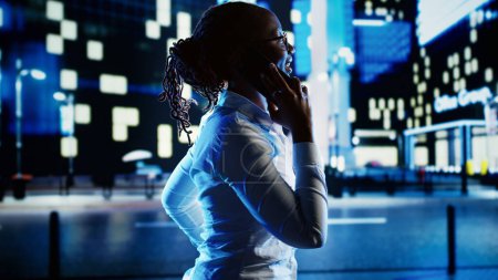 Téléchargez les photos : Femme afro-américaine flânant dans le paysage urbain la nuit, profitant d'une conversation au téléphone avec son mari. Femme d'affaires marchant à la maison du travail ayant un appel téléphonique pour faire le trajet aller plus vite - en image libre de droit