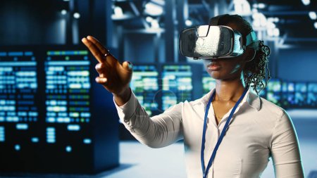Photo for African american certified developer immersed in virtual reality at data center, doing units maintenance. Licensed technician using VR headset to optimize servers performance, checking operations - Royalty Free Image