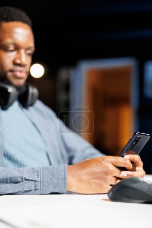 Foto de Afroamericano chico mensajes de texto en la aplicación de teléfono, trabajando desde casa en su estación de trabajo. Estudiante freelancer masculino revisando sitios web y escuchando el programa de entrenamiento en línea, e learning. - Imagen libre de derechos