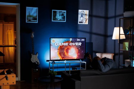 Photo for Player is dissatisfied after failing to win shooter tournament while engaging in web based multiplayer battle at night. Unhappy adult playing first person online action games on home television. - Royalty Free Image