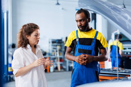 Freundlicher Ingenieur hilft dem Kunden bei der Autoversicherung in der Autowerkstatt. Werkstattarbeiter schaut mit Frau über Autoteile, repariert ihren defekten Kraftstofftank während der Inspektion