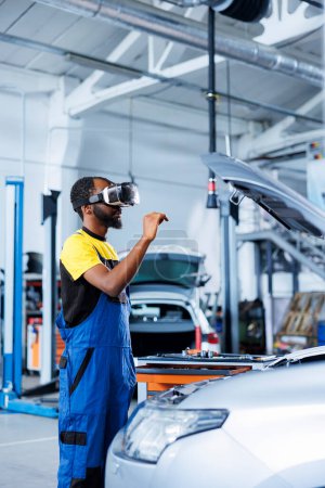Repairman in auto repair shop using virtual reality to visualize car fuel tank in order to fix it. BIPOC adept garage worker wearing futuristic vr headset while working on busted vehicle
