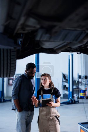 Fachkundiger Mechaniker in der Garage mit Laptop, um Checkliste zu folgen, während Wartung am Auto, im Gespräch mit dem Kunden. Spezialist in der Kfz-Werkstatt checkt Autoreifen neben dem Kunden