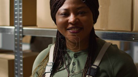 Portrait eines fröhlichen afrikanisch-amerikanischen Lagerleiters, der online aktualisierte Produktinfos eingibt, zwischen Verkaufsregalschränken steht und Pakete identifiziert, die versandbereit sind, Nahaufnahme