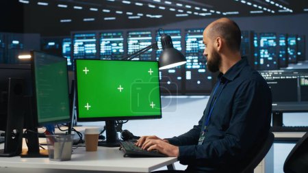Hombre escribiendo código en la computadora de pantalla verde en el centro de datos de alta tecnología con filas de servidores que proporcionan recursos informáticos. Especialista en TI que utiliza una maqueta de PC para supervisar los montajes en rack de datos operativos