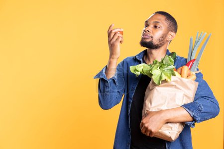 Afrikanisch-amerikanischer Typ bewundert eine Tomate und riecht das frische Aroma vor gelbem Hintergrund, er trägt eine Papiertüte mit Bio-Lebensmitteln. Glücklicher Mensch mit ethisch vertretbarem Gemüse zufrieden.