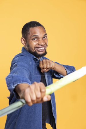 Photo for African american adult mimicking a combat move with a leek, holding produce as a sword against yellow background. Confident silly guy acting silly with locally grown green onion. - Royalty Free Image