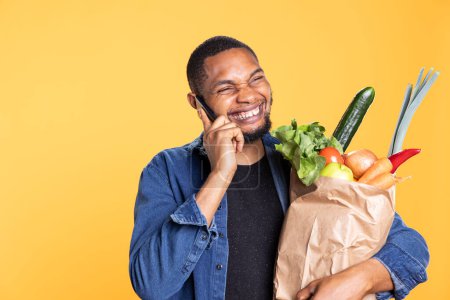 Photo for African american excited guy chatting on phone call and holds bag of groceries, discussing with his friends on remote connection line. and Carrying ethically sourced produce in studio. - Royalty Free Image