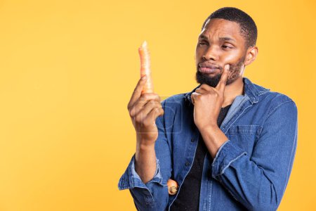 Pensive african american man thinking what recipe to cook with a carrot, holding freshly harvested vegetable for healthy eating concept. Confident person examining organic ripe veggies.