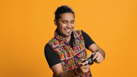 Happy gamer celebrating after winning game on gaming console, studio background. Delighted Indian man bragging after being victorious in videogame, defeating all enemies using gamepad, camera A