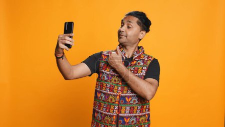 Foto de Un narcisista indio usando un celular para tomar selfies desde todos los ángulos. Vano usuario de medios sociales tomando fotos usando la cámara selfie del teléfono, sonriendo felizmente, fondo del estudio, cámara B - Imagen libre de derechos