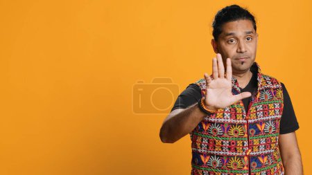 Stern indian man doing stop hand gesture sign, complaining. Authoritative person doing firm halt sign gesturing, wishing to end concept, isolated over studio background, camera A
