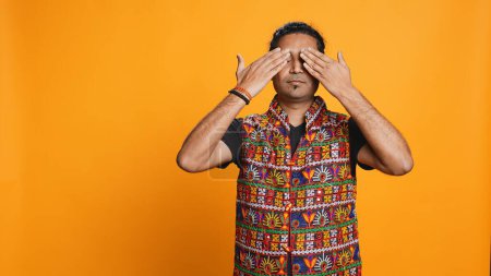 Man wearing traditional indian clothing covering eyes, ears and mouth, imitating three wise monkeys, doing dont see, dont hear and dont speak hand gesturing concept, studio background, camera B