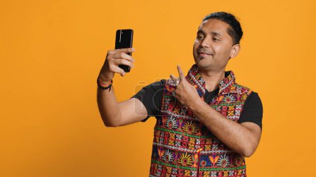 Foto de Un narcisista indio usando un celular para tomar selfies desde todos los ángulos. Vano usuario de medios sociales tomando fotos usando la cámara selfie del teléfono, sonriendo felizmente, fondo del estudio, cámara A - Imagen libre de derechos