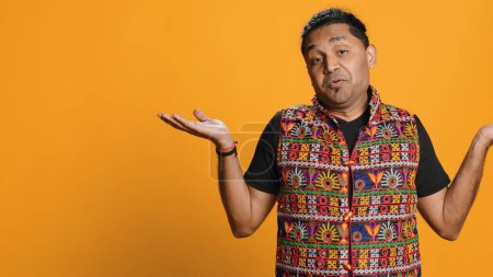 Indian man shrugging shoulders, unable to provide answer, having detached apathy facial expression. Apathetic person doing hand gesturing showing lack of knowledge, studio background, camera A