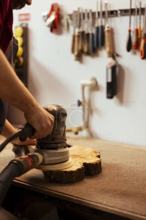 Photo for Cabinetmaker at workbench using orbital sander with fine sandpaper on lumber to achieve smooth finish. Woodworking specialist in assembly shop uses angle grinder on wood for professional results - Royalty Free Image