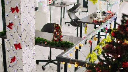 Zoom in on beautifully decorated Christmas pine tree placed in empty office. Xmas tree adorn with red bows and baubles in festive workplace filled with lights during winter holiday season