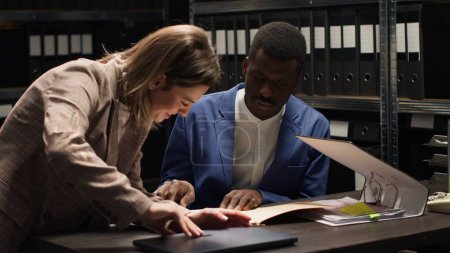 Photo for African american detective holding case files enters office with caucasian policewoman carrying backpack. Policeman discussing clues and evidence while female investigator places laptop on desk. - Royalty Free Image