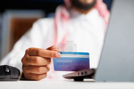 Empresario árabe enfocado en un escritorio moderno, absorto en un eficiente trabajo en línea. Usando tecnología inalámbrica, escribe, investiga y compra de forma segura con una tarjeta de crédito.