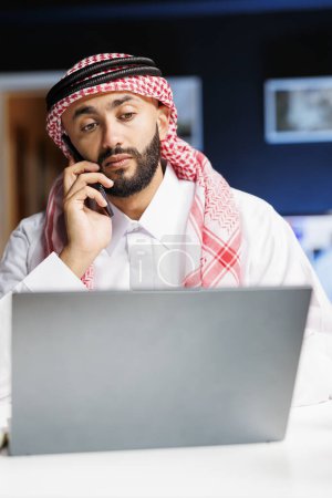 Imagen detallada del hombre árabe utilizando un ordenador portátil y un dispositivo móvil para la conversación y el estudio, demostrando la aptitud tecnológica. Multitarea chico musulmán conversando en su móvil e intercambiando notas.