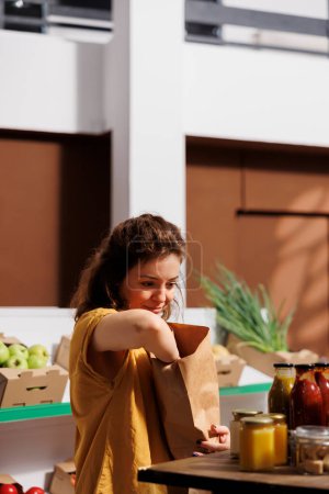Foto de Smiling woman buying bio produce in zero waste store using reusable biodegradable paper bags to minimize plastic usage. Green living customer looking for ethically sourced pantry staples - Imagen libre de derechos
