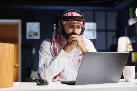 Un hombre enfocado en vestimenta árabe tradicional trabaja diligentemente en su escritorio, utilizando tecnología inalámbrica. Investiga en línea, se comunica y participa en videoconferencias en su computadora personal.