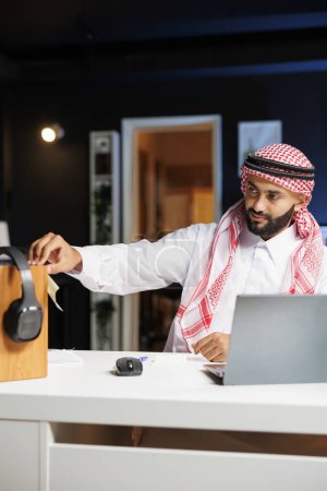 L'homme du Moyen-Orient traditionnellement vêtu travaille sur son ordinateur portable et prend des notes alors qu'il est assis dans son bureau. Jeune musulman plaçant une note collante sur une planche en bois avec écouteurs.