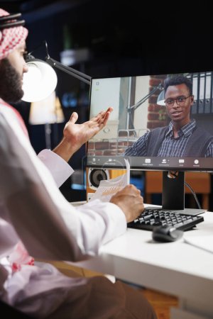 La videollamada aparece prominentemente en la pantalla de la computadora, mostrando la conversación entre el freelancer musulmán y el colega afroamericano. La imagen subraya el uso de la tecnología moderna por parte de los jóvenes.