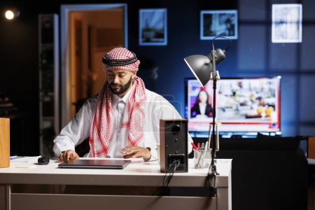 Hombre de Oriente Medio en ropa árabe sentado a la mesa con su portátil digital preparándose para el trabajo. Hombre musulmán dedicado está a punto de abrir su computadora inalámbrica para navegar por Internet.