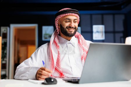 Smiling Arab guy works diligently at office desk, engages in digital tasks and writes on his notepad with laptop nearby. He efficiently conducts online research, embodying dedication and productivity.