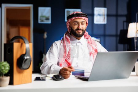 Hombre de negocios árabe en la oficina del hogar, navegar eficientemente y escribir en su computadora portátil. Él multitarea con la investigación en línea, compras, y los pagos con tarjetas de crédito y débito.