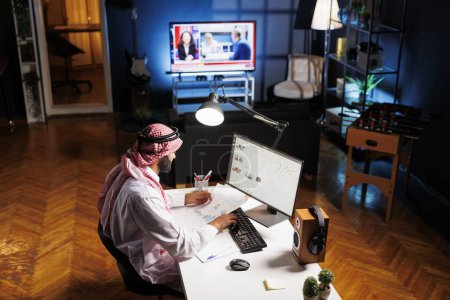 Muslim guy with paperwork diligently conducting online tasks efficiently. Young man in Islamic clothing manages his work with dedication and meticulousness by using a desktop PC. Overhead shot.