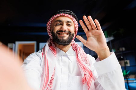 Pov von einem fröhlichen muslimischen Kerl in traditioneller Kleidung, der Videoanrufe tätigt. Lächelnder Araber mit Kopftuch greift nach digitalem Gerät und winkt in die Kamera, macht Online-Vlogging.