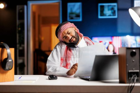Traditionell gekleideter muslimischer Mann sitzt an seinem Schreibtisch und unterhält sich auf seinem Smartphone, während er Papiere in der Hand hält. Araber greift nach Forschungspapieren, während er auf seinem Handy spricht.