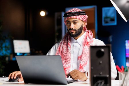 Arabischer Geschäftsmann tippt selbstbewusst auf einem Laptop an einem modernen Schreibtisch. Männliche muslimische Person, die ihren PC für E-Mails und Surfen in einer ruhigen und professionellen Arbeitsumgebung nutzt.