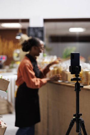 Black woman at local supermarket, promoting healthy and sustainable living. A smartphone is used to shoot a vlog to highlight eco friendly solutions, fresh vegetables and glass jars. Foreground focus.