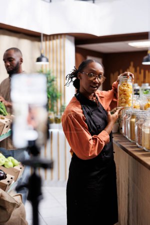Joven mujer negra graba un video en una tienda de basura cero promoviendo productos frescos y orgánicos. Utiliza un soporte de trípode mientras exhibe envases sostenibles y opciones saludables.