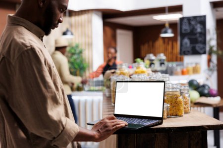 Un supermercado ecológico utiliza un dispositivo digital para mostrar sus productos. Cliente masculino afroamericano con una computadora portátil que muestra una pantalla blanca aislada está buscando artículos de bio-alimentos.