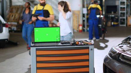 Green-Screen-Laptop auf Arbeitstisch in belebter Garage neben professionellem Werkzeug platziert, während Mitarbeiter im verschwommenen Hintergrund herumstreifen. Chroma-Schlüsselgerät in der Autowerkstatt