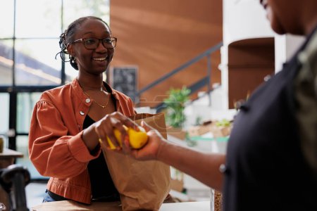 Closeup shows two African American women holding freshly harvested lemons above the checkout counter. The image focused on female customer giving fruits to black vendor for weighing.