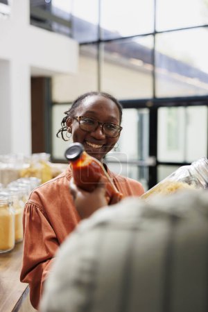 Eco friendly African American farmer sells bottled organic products at local market. Smiling black female client listening to vendor about freshly harvested and sustainable options in store.