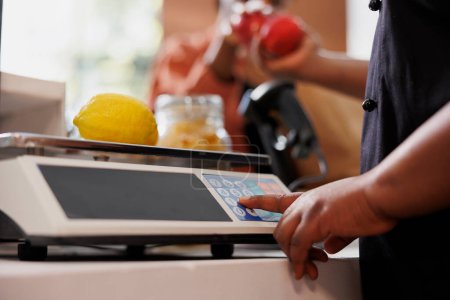 Close-up shot of a pair of African American hands holding red apples over the checkout counter. Selective focus on a female vendor weighing locally grown lemon on a scale.
