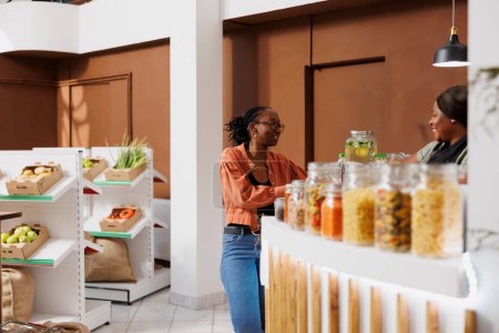 La cajera negra sirve a un cliente en una tienda de comestibles ecológica, ofreciendo alimentos recién cosechados y sostenibles. Cliente afroamericano hablando con el vendedor en el mostrador de pago.