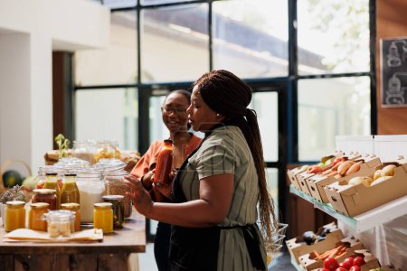 Schwarze Frau mit Brille beim Einkaufen im Zero-Waste-Laden auf der Suche nach landwirtschaftlichen Massenprodukten. Kunde kauft Grundnahrungsmittel im örtlichen Lebensmittelgeschäft mit Hilfe einer Verkäuferin.