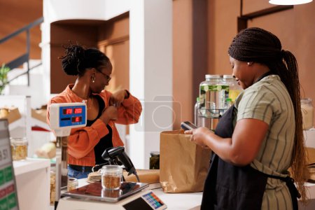 Après avoir payé pour des produits bio qu'elle a sélectionnés dans un magasin respectueux de l'environnement, la femme noire remet sa carte de crédit dans sa poche. Au comptoir de caisse, marchand et client effectuent une transaction sans espèces.
