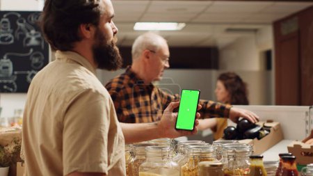 Foto de Hombre usando el teléfono inteligente de pantalla verde en el supermercado de cero residuos para comprobar los ingredientes para una receta saludable. Cliente en la tienda de comestibles local utiliza el teléfono clave de croma mientras que las compras de verduras orgánicas - Imagen libre de derechos