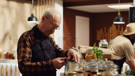 Portrait of smiling old zero waste vendor properly sealing bulk items decomposable glass containers. Elderly man in environmentally friendly local grocery shop arranging merchandise