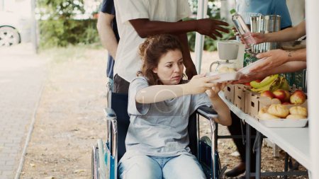 Foto de Mujer caucásica en silla de ruedas recibiendo comida gratis de trabajadores de caridad en el programa sin fines de lucro al aire libre. Equipo de voluntarios de socorro del hambre repartiendo provisiones a personas necesitadas sin hogar y discapacitadas. - Imagen libre de derechos