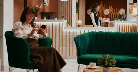 Alte Frau liest SMS nach internationalem Flug und wartet im Fünf-Sterne-Resort auf den Check-in. Senior Tourist surft Smartphone-App in Lounge-Bereich, entspannen in luxuriöser Lobby.